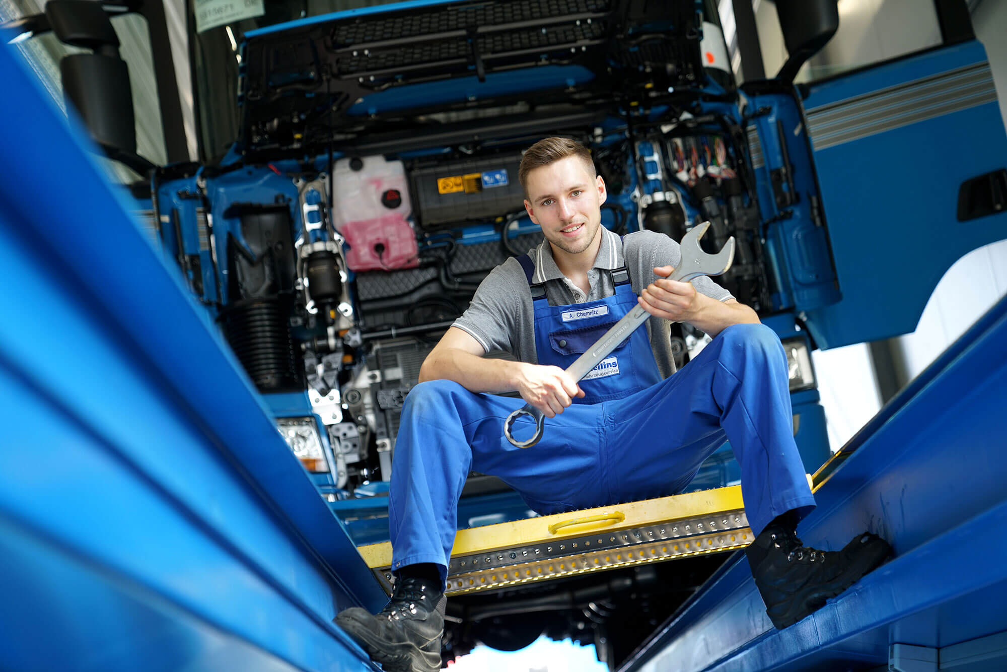 Ein junger Mann in seiner Ausbildung bei Reilling als Mechatroniker für LKWs. Mit einem Schraubenschlüssel in der Hand sitzt er lächelnd an einer LKW-Fahrzeuggrube. Im Hintergrund ist ein großer LKW mit geöffneter Front zu erkennen, bereit, von ihm repariert zu werden.