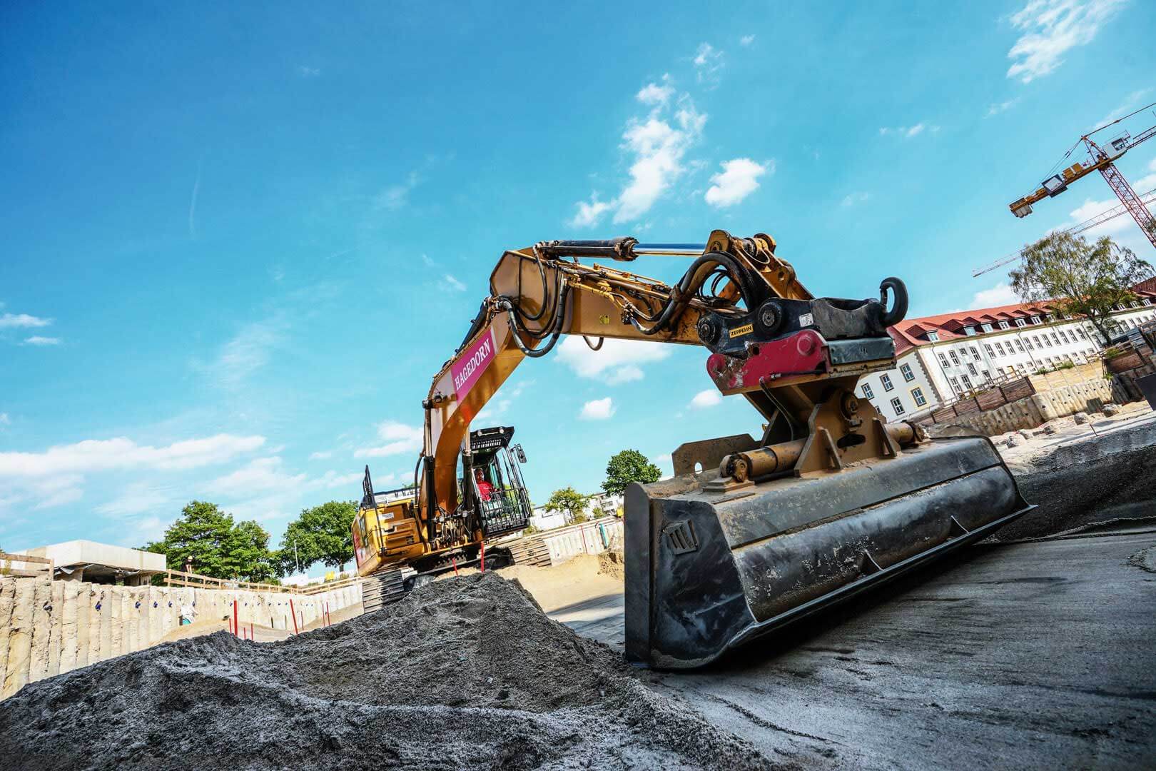 Eine eindrucksvolle Baustellenszene: Der gewaltige LongFrontBagger von Hagedorn steht im Vordergrund, arbeitend auf einer Baustelle. Seine Schaufel begradigt den Boden.