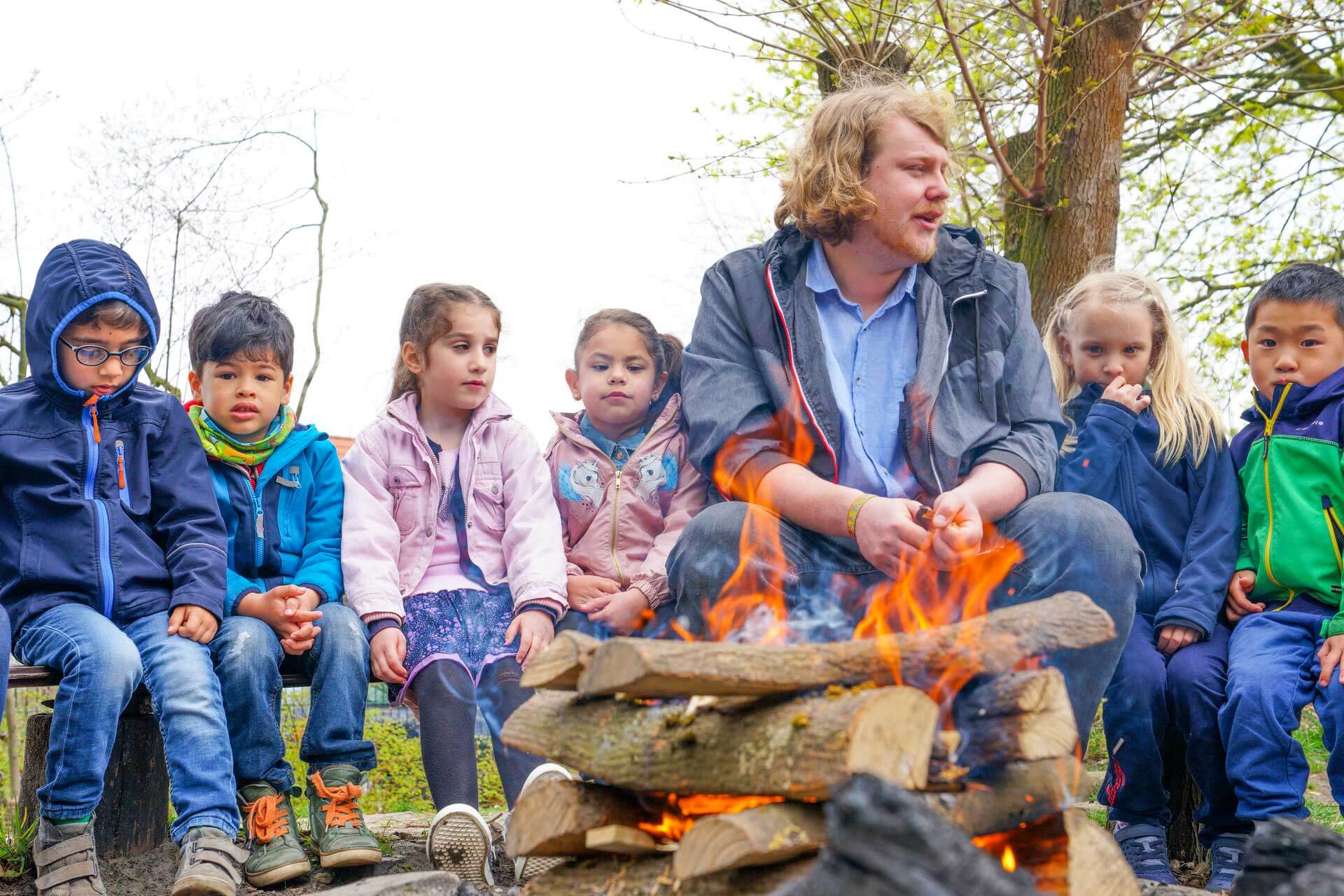 Ein Kindergärtner und fröhliche Kinder sitzen gemeinsam tagsüber um ein Lagerfeuer.