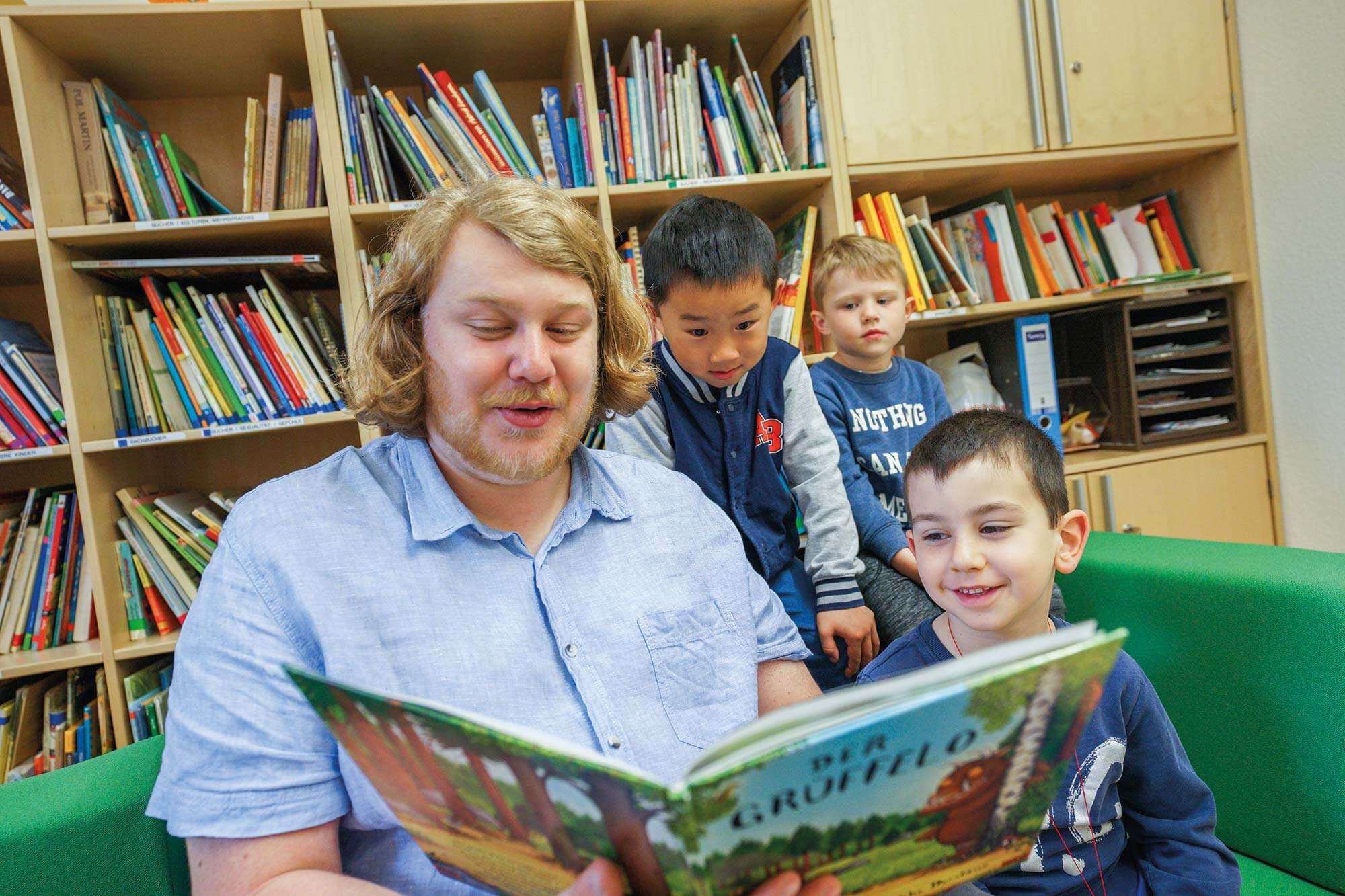 "Ein junger Mitarbeiter der Städtischen Kindertagesstätte liest begeistert aus einem Kinderbuch vor, während drei aufmerksame Kinder gespannt lauschen
